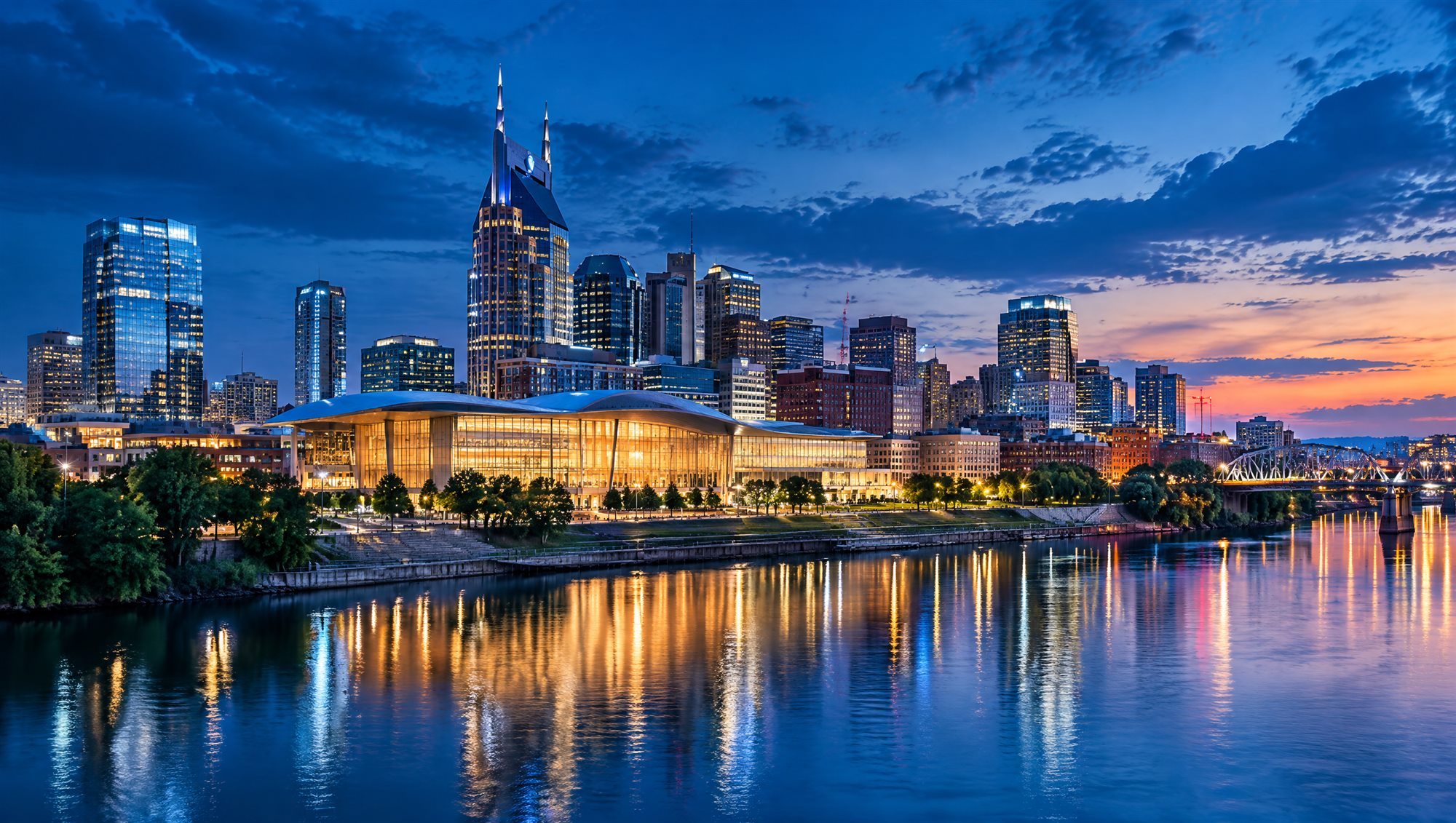 The Nashville skyline at dusk across the river, with a convention hall glowing on the bank
